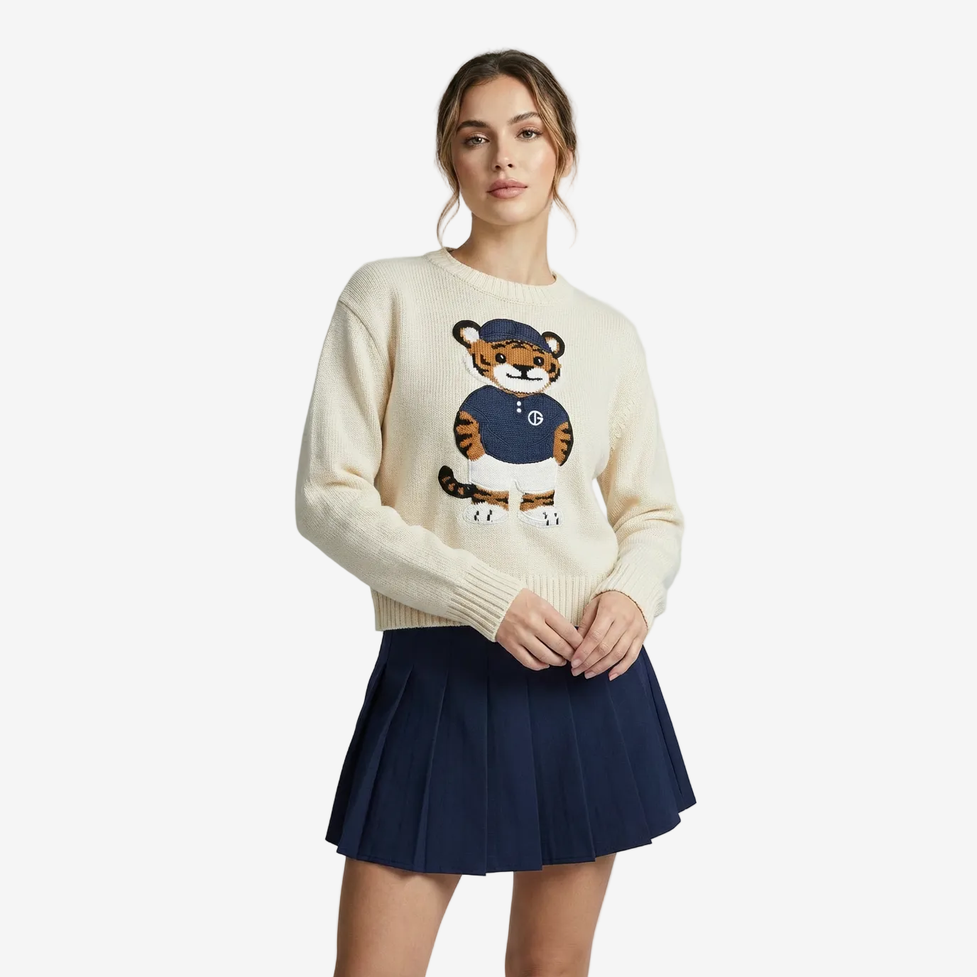 Woman wearing a cream sweater with a tiger design and a navy pleated skirt on a light gray background