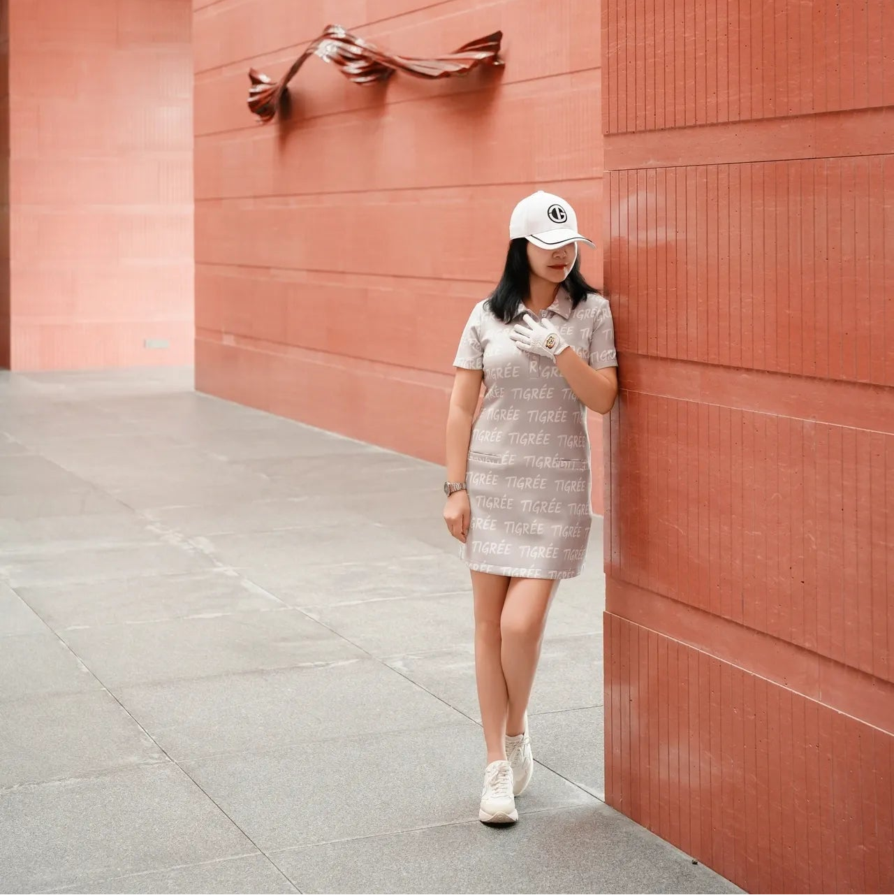 Woman in a dress and cap standing against a pink wall with a sculpture.